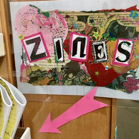 Sign reading "Zines" with an arrow pointing to a bookshelf.