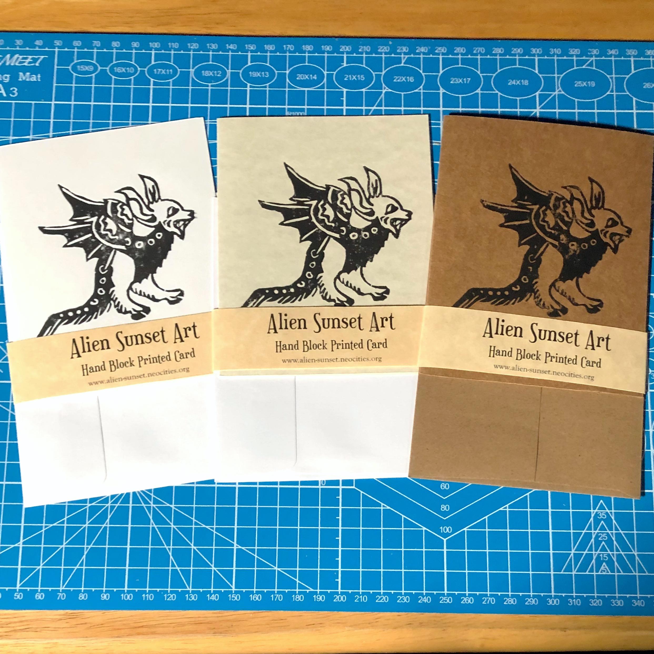 a set of three cards laid out on a blue cutting mat. a brown Kraft card, a plain white card, and a cream parchment pattered card. each card has a yellow parchment strip of paper wrapped around it that says "Alien Sunset Art" Hand Block Printed Card.

the block print on the cards is of  a small lizard creature with leathery wings and a devils face