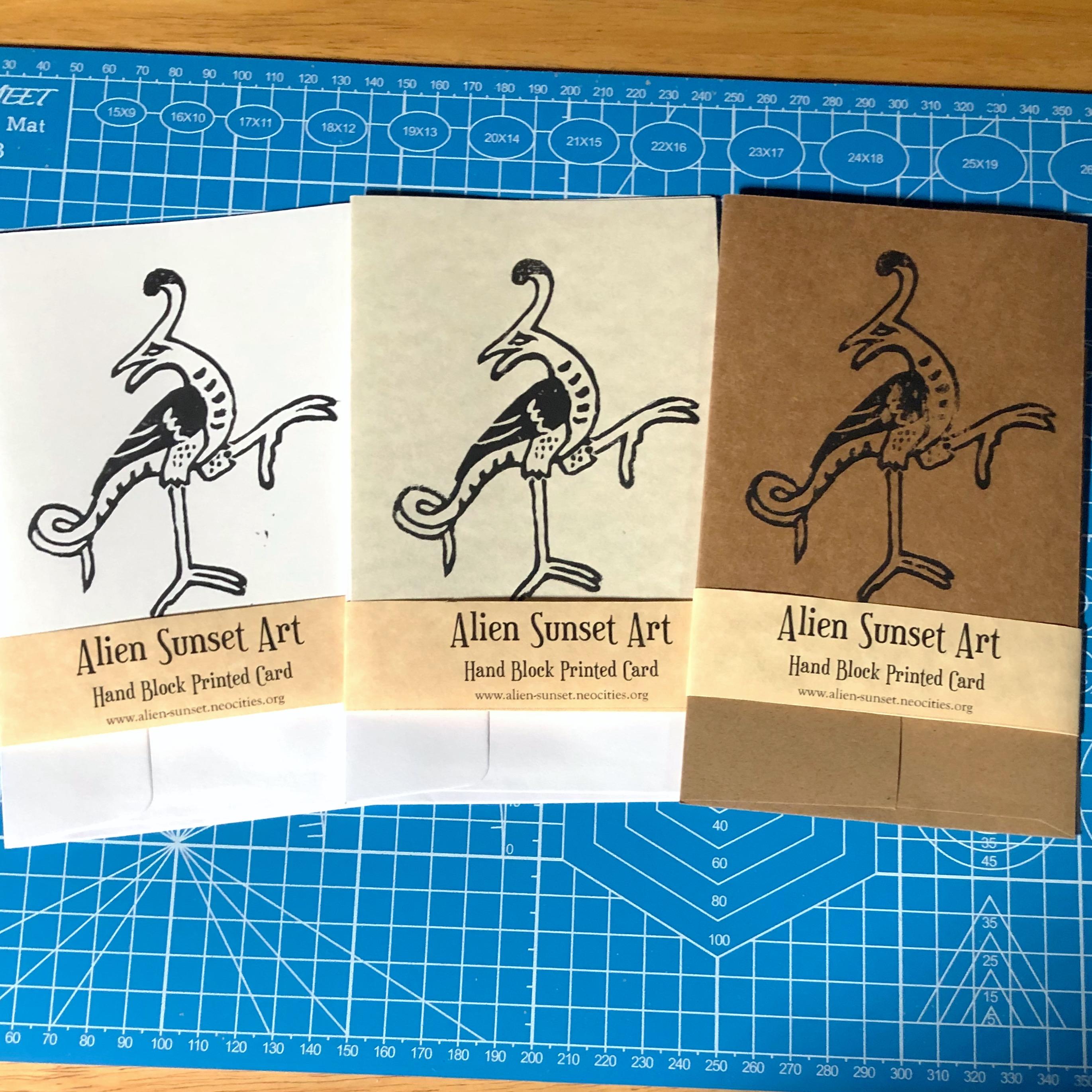 a set of three cards laid out on a blue cutting mat. a brown Kraft card, a plain white card, and a cream parchment pattered card. each card has a yellow parchment strip of paper wrapped around it that says "Alien Sunset Art" Hand Block Printed Card.

the block print on the cards is of  a  tall stork like creature with a lizards tail with one leg lifted as if to grab something