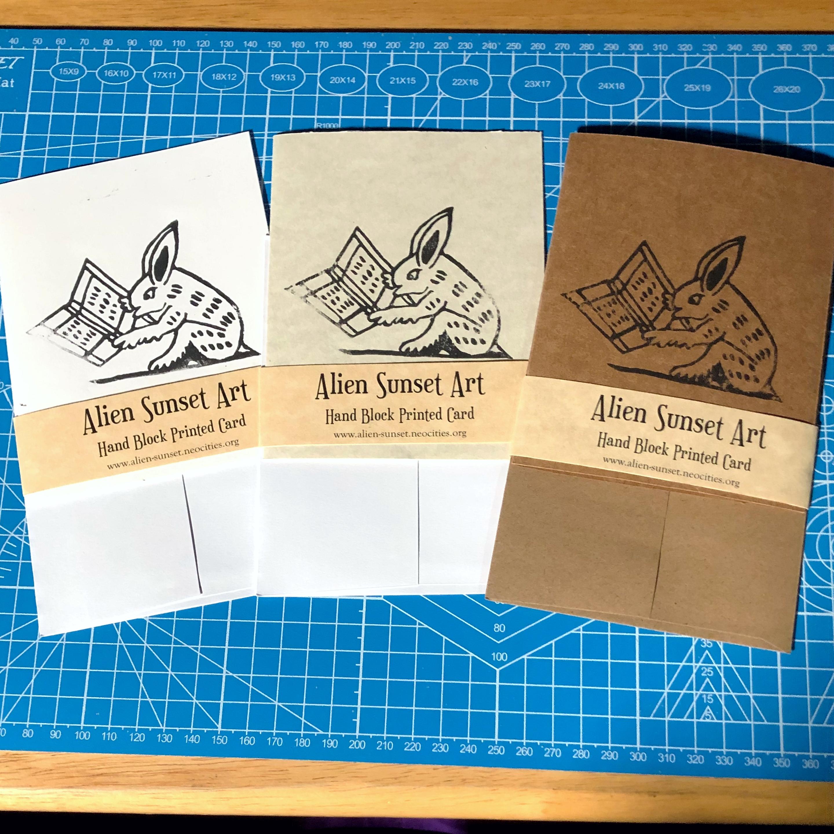 a set of three cards laid out on a blue cutting mat. a brown Kraft card, a plain white card, and a cream parchment pattered card. each card has a yellow parchment strip of paper wrapped around it that says "Alien Sunset Art" Hand Block Printed Card.

the block print on the cards is of  a bunny reading a book