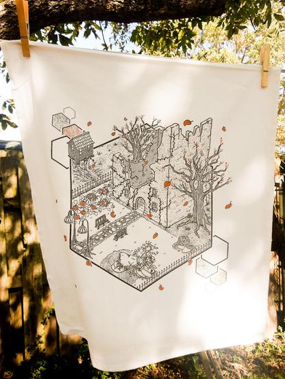a white tea towel hanging from a clothesline with wooden clothespins. The towel is printed with an image in black isometric linework with orange accents, depicting a scene of the Baba Yaga running out of the courtyard of a crumbling building. There's a pumpkin patch, crumbling statue, ivies and trees. 