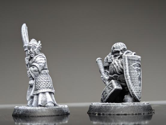 Two unpainted fantasy miniatures stand side by side: a robed warrior gripping a raised glaive and a stocky fighter with a hammer and shield, both captured in a stark, high-contrast grey.
