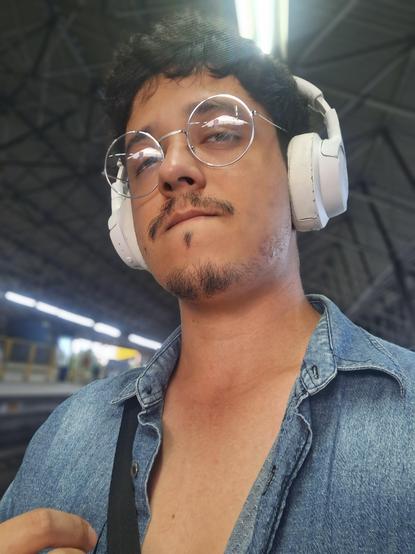 Eu (homem, pele parda, cabelo, bigode e barbicha pretos, vestindo óculos prateados, fones de ouvido brancos, uma alça de bolsa preta e uma camisa jeans desabotoada), numa estação de trem, olhando para a câmera