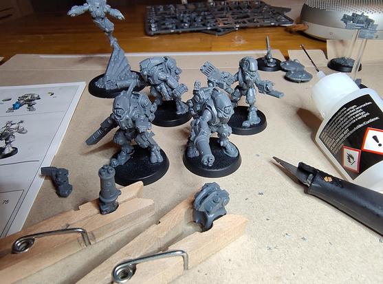 Four of five XV26 stealth battlesuits assembled but unpainted on a table. Some parts for the fifth in the front. Building instructions, glue and a knife frames the picture. A half-assembled Shadowsun model in the background.