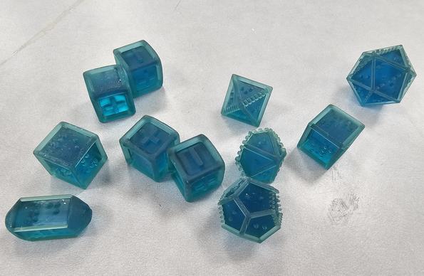 A set of polyhedral dice in translucent blue resin, with Braille markings for the numbers. The dice set includes an oblong style 4-sided die, 2 six-sided dice, an 8-sided die, 10-sided die, 12-sided die, and 20-sided die. A set of 4 Fudge or Fate dice completes the set. The braille dice have a small ridge to indicate the lower edge of each dice face.