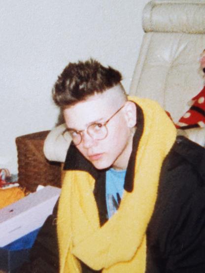 Portrait of a teenage person wearing round-framed glasses, a black trenchcoat, a long yellow knit scarf, with spiky hair shaved on the sides.