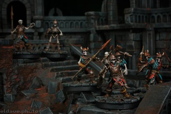 Eight skeletons, wearing ragged clothing and carrying rusted weapons and armor walking through a ruined stone city. Models prepped for games of Frostgrave.