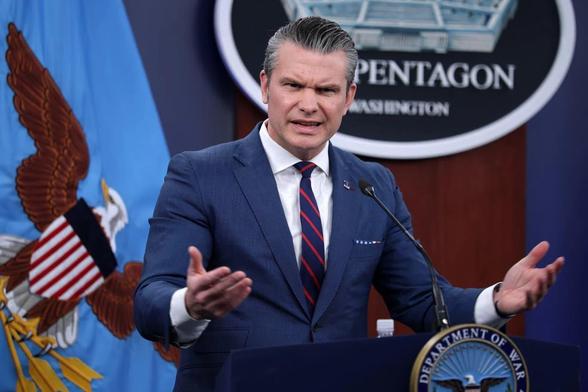 US Defense Secretary Pete Hegseth at the podium (erroneously labelled “Department of War”) of a Pentagon press conference. He holds his hands, palm up, in a motion of supplication; his face suggests he’s trying to crap out a particularly uncooperative ferret.