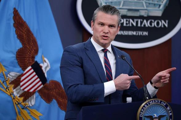 US Defense Secretary Pete Hegseth at the podium (erroneously labelled “Department of War”) of a Pentagon press conference. His expression suggests that the aforementioned ferret has slipped back in. Both hands point off-camera, as if trying to distract the audience so he can deal with the problem manually.