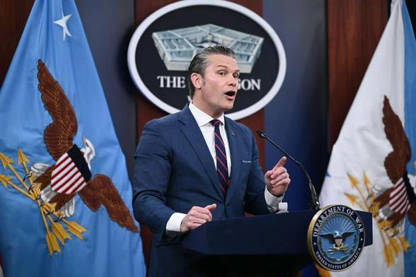 US Defense Secretary Pete Hegseth at the podium (erroneously labelled “Department of War”) of a Pentagon press conference. One hand rests on the podium, and the he points with the other’s index finger as if he has something particularly relevant to say. His expression suggests he’s channelling the spirit of a long-discarded sex doll.