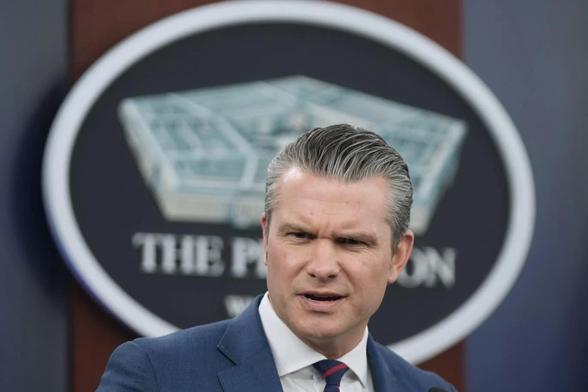 US Defense Secretary Pete Hegseth at the podium (erroneously labelled “Department of War”) of a Pentagon press conference. His expression suggests he’s seen his tiny, unmanly tie knot in a reflective surface, and can’t believe his minders let him out in public that day.