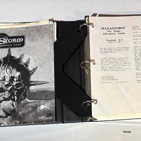 A three-ring binder containing the playtest manuscript for "ManaStorm: The Magic Adventure Game," featuring a black and white cover illustration of a character with a spiked helmet and a weapon. The title "MANASTORM The Magic Adventure Game" is prominently displayed at the top, along with the version number and date on the first page inside the binder. Additional pages include loose character sheets and player handouts, indicating the game's mechanics and spells.