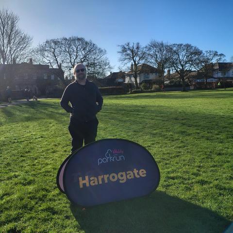 A man dressed in black running gear and sunglasses, standing on grass, behind a sign saying "parkrun Harrogate". Behind him are trees and houses.