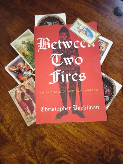 Paperback copy of Between Two Fires surrounded by catholic prayer cards.