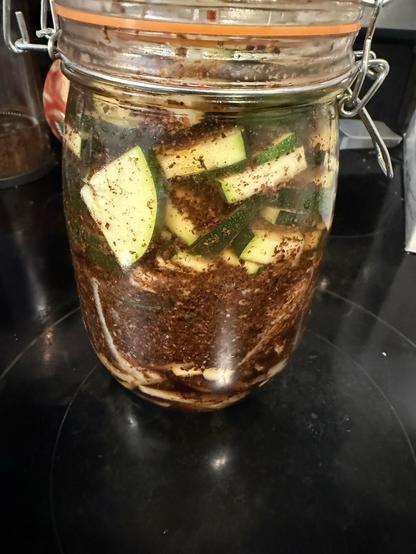 A clear jar filled with sliced zucchini submerged in a brown, spicy marinade. The jar is sealed and placed on a dark countertop.