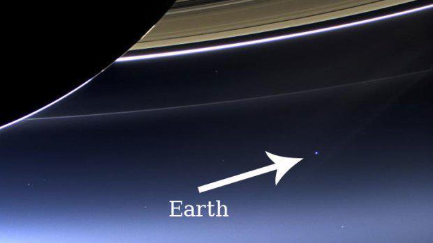 Photo from the Cassini probe showing part of Saturn in the upper left corner, an ominous dark portion of a sphere, with a few striated rings aross the top of the shot, with a tiny speck of light in the bleak, dark distance. A white arrow points to the insignificant dot with the label "Earth."