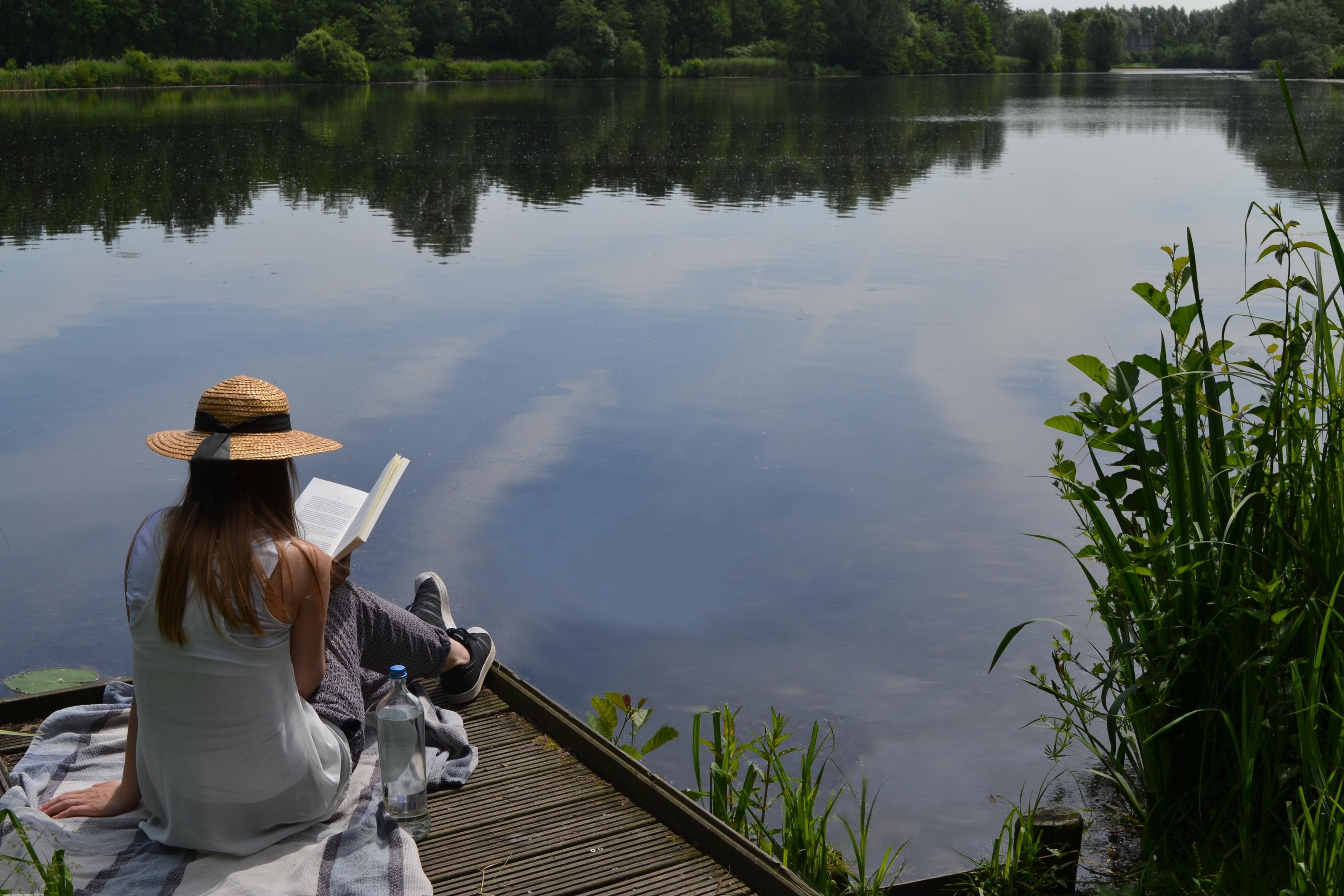 Scene of a woodland lake, still waters reflecting the terrain, with a woman ina  wide-brimmed sun hat sitting on a pier reading a book.
