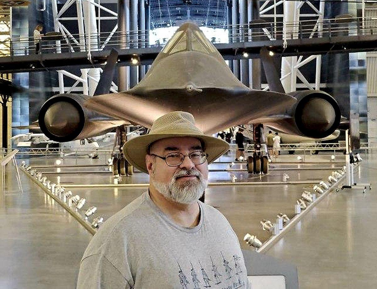 Doofy looking old white guy in beard, glasses, and a brimmed hat standing centered on the front of an SR-71 Blackbird reconnaissance aircraft, a triangular cockpit above his head flanked by two massive black jet engines.

https://airandspace.si.edu/collection-objects/lockheed-sr-71-blackbird/nasm_A19920072000