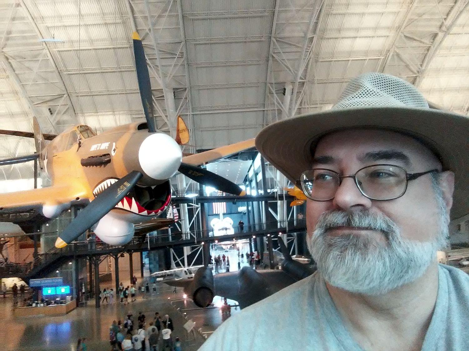 Doofy looking old white guy in beard, glasses, and a brimmed hat on the right looking to the left into the front of a shark-mouthed Curtiss P-40E Warhawk, a brown WWII fighter plane hanging from the museum’s hangar ceiling.

https://airandspace.si.edu/collection-objects/curtiss-p-40e-warhawk-kittyhawk-ia/nasm_A19650242000