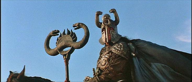 A still from Conan the Barbarian (1982): an armoured rider in a helmet with a moon and sun on its brow, and two serpents facing each other like horns, bears the twin-headed serpent standard of Thulsa Doom.