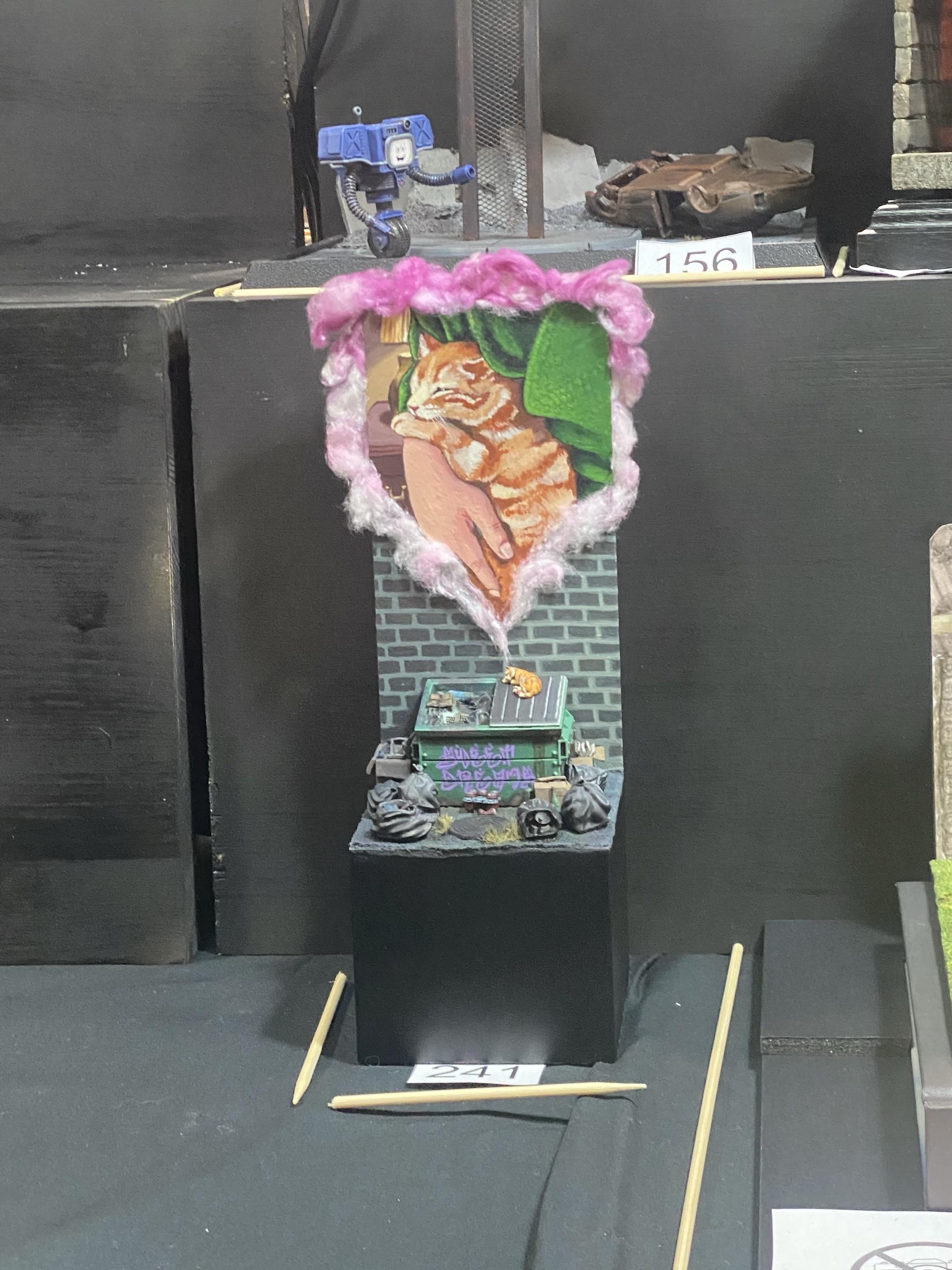 Diorama of a cat sleeping on top of a dumpster surrounded by trash bags and boxes. The cat has a thought bubble dreaming of itself sleeping in the crook of a person's arm. The presumed title of the piece is "sweet dreams", written in graffiti lettering across the dumpster.