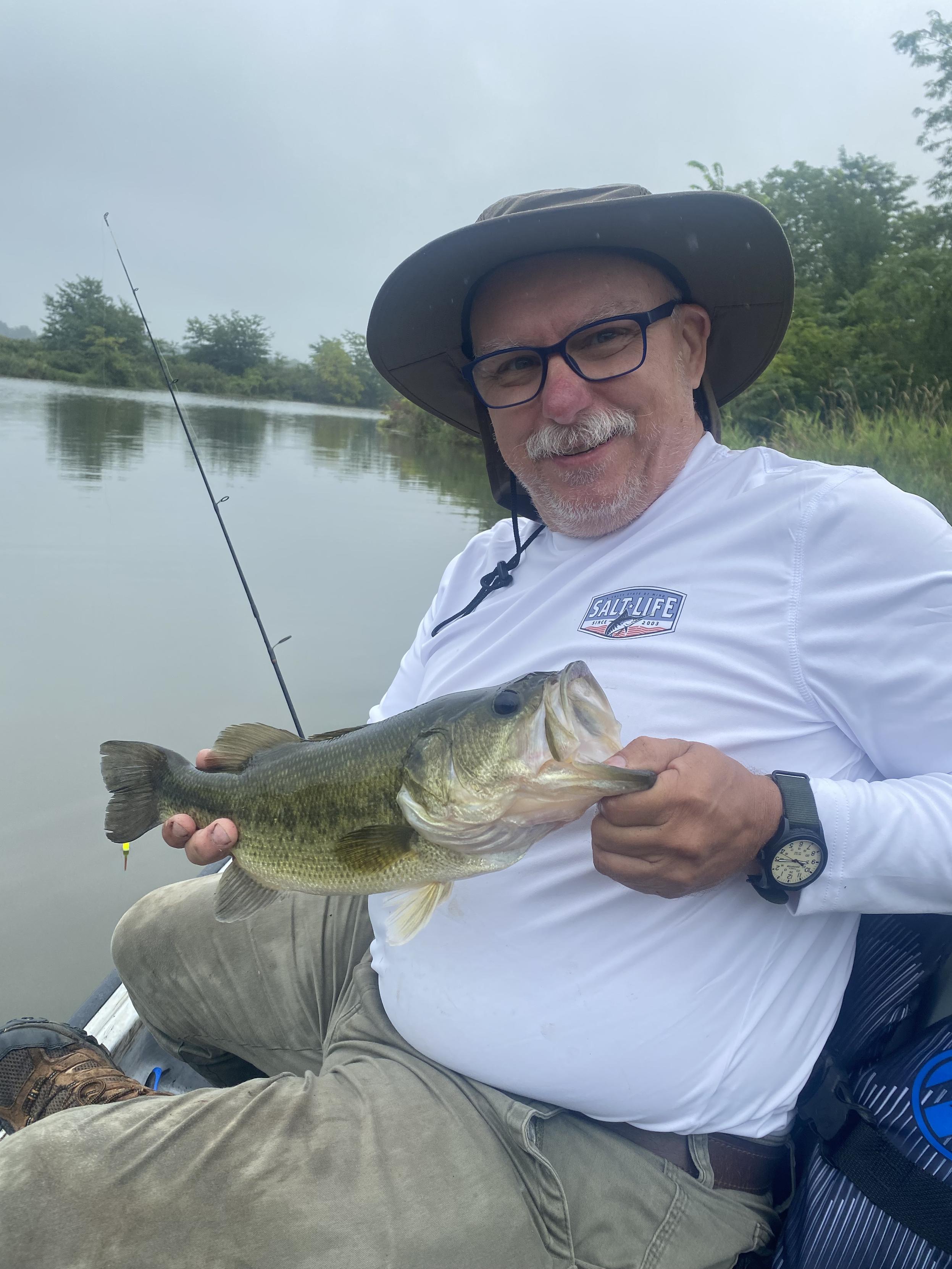 A man in a white long sleeve fishing shirt, wearing glasses and a sun hat, holds a largemouth bass fish that he's just caught.