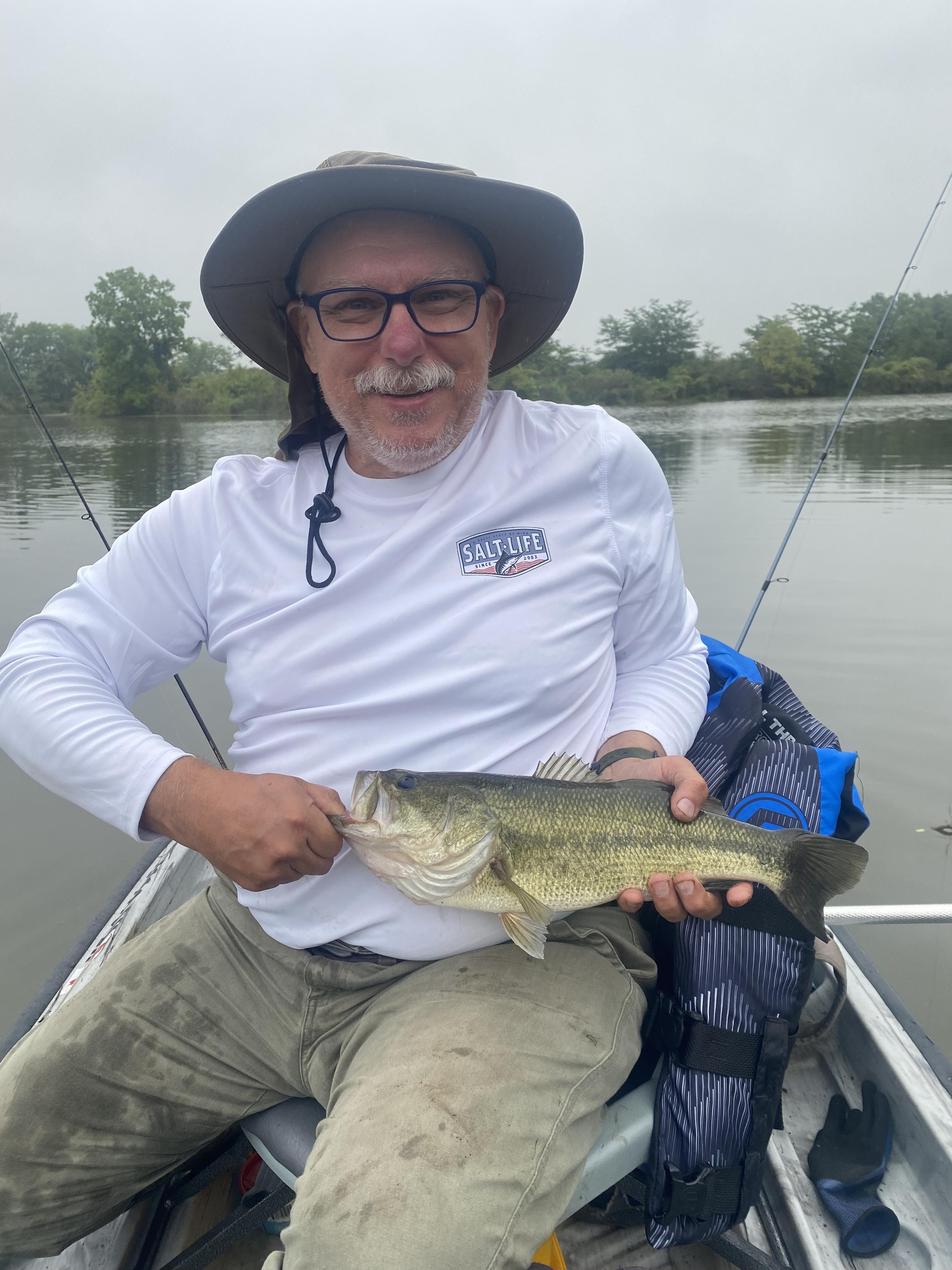 A man in a white long sleeve fishing shirt, wearing glasses and a sun hat, holds a largemouth bass fish that he's just caught.