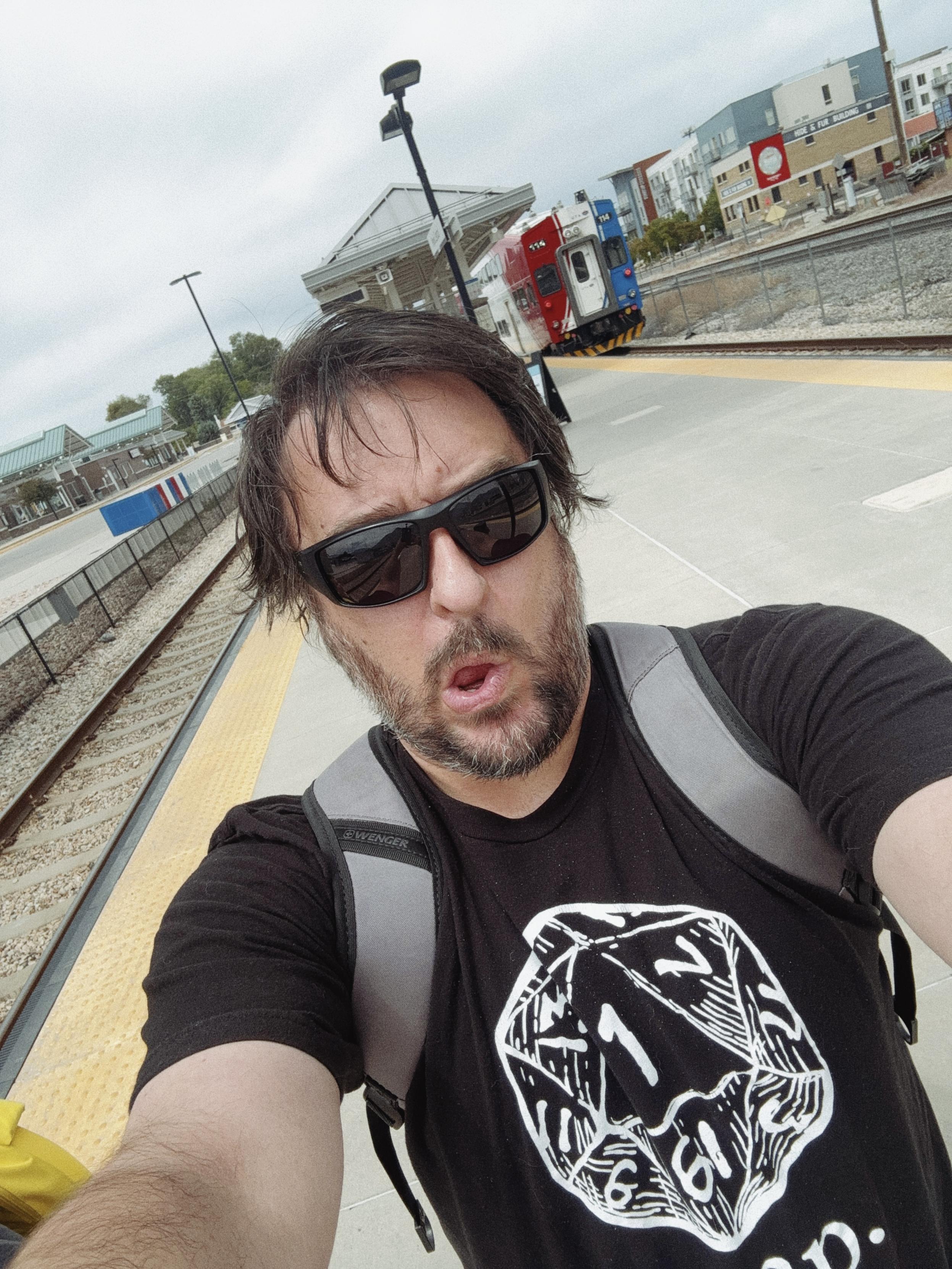 Selfie of a guy with shaggy hear and short beard, wearing sunglasses, a backpack, and a black t-shirt with a 20-sided die that rolled a 1 and says "crap" below it. He's walking along a train platform in Provo, Utah. This picture was taken on the way to Conclash, formerly known as Dead Wars, the Guinness World Record-holder for the largest game of Dungeons & Dragons ever played at 1,227 people.