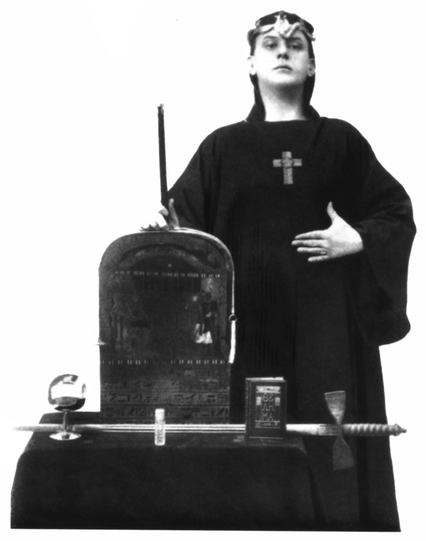 A black-and-white photo of occultist Aleister Crowley from his 1912 treatise on ceremonial magick, titled “Magick” (alternatively, “Book Four” or “Liber ABA”). Crowley stands tall, looking directly at the viewer, and wearing an Egyptian-inspired headpiece, resembling a keffiyeh with a sculpted cobra sitting above his brow. He bears a cross amulet over his black robe; his left hand rests on his ribs, and his right hand—holding a black wand erect—rests atop an upright stone tablet. On the same, c…
