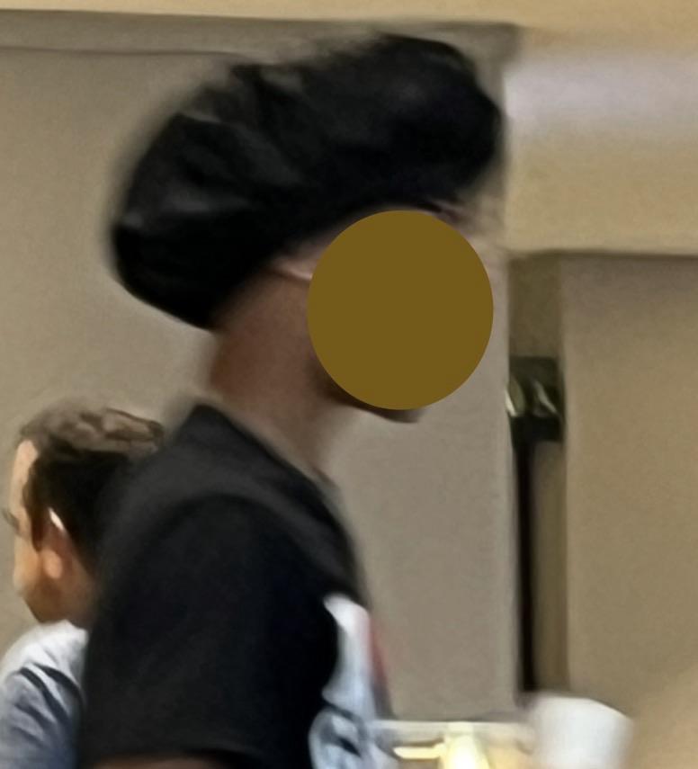 A person is seen in profile wearing a black head wrap and a black shirt. The background is blurred, with another individual partially visible.