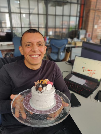 Chris sitting in front of his computer, holding a cake decorated with cream, Oreos, pretzels etc.