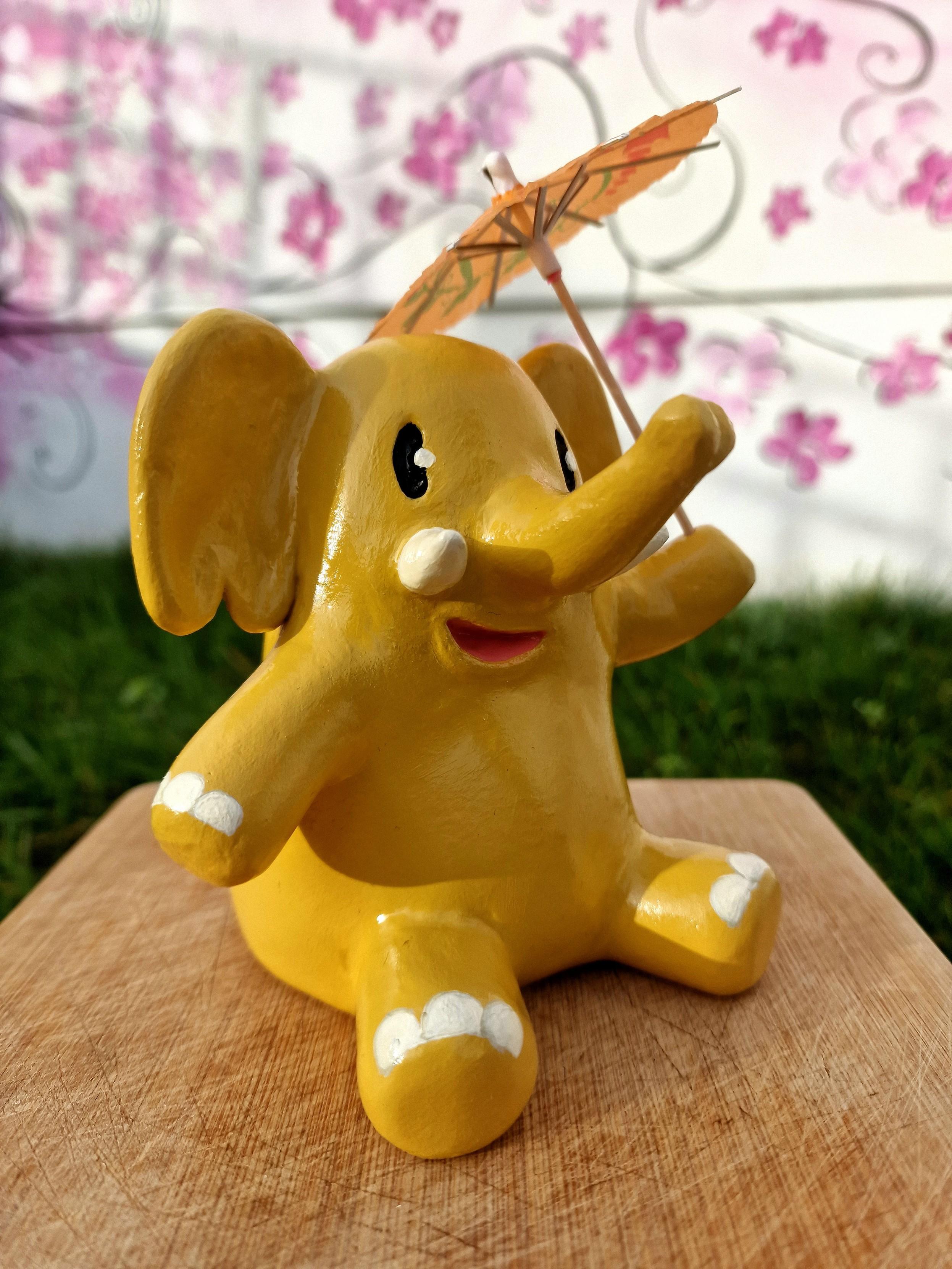 Clay ornament of a yellow elephant, the mastodon mascot, sitting down and holding up a cocktail umbrella.