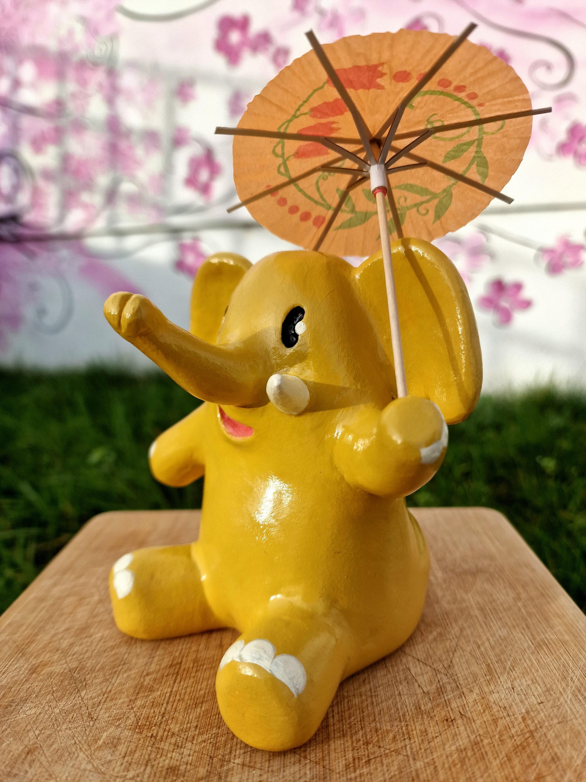 Clay ornament of a yellow elephant, the mastodon mascot, sitting down and holding up a cocktail umbrella.