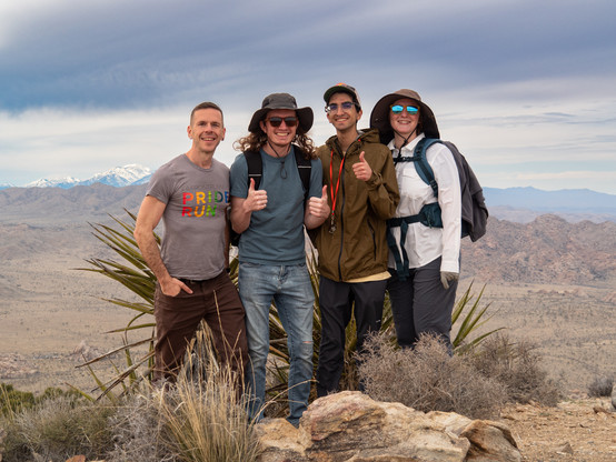 Four people in hiking gear pose for a photo on the summit of a mountain, with a desert landscape visible below