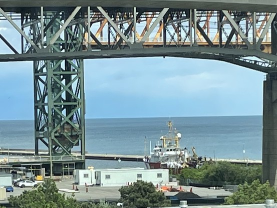 Photo of a red and orange ship exiting a canal under an orange lift bridge with a large lake in the background. CCGS Limnos under Burlington Lift Bridge. Photo from DFO.