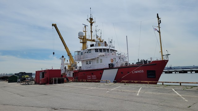 Photo of a red ship with a white superstructure and white slash on the front and Limnos on the front. Coast Guard and Canada are visible on the side. A large yellow onboard crane is loading equipment. Photo from DFO.