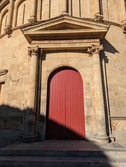 Un plano muy cercano de un edificio histórico, construido con bloques de arenisca, lo que le da un aspecto cálido a la piedra.

La puerta está hecha en tablones de madera y pintada de color rojo. Está adornada con dos columnas neoclásicas y un frontón triangular.
Recuerda mucho a edificios de la antigüedad romana, y la frase en latín sobre la puerta acentúa esto.

Está escrita con letras grandes y rectas, en un color rojizo, similar al de la arcilla. Es la cita del pie de foto.