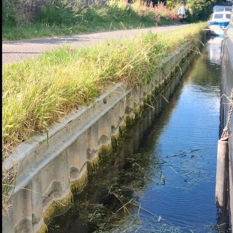 Photo looking at the water between the bank of the canal and the entire length of the boat. The distance is about 1 meter. On the bank the grass is mowed and there's a towpath. The bank itself is steel piling. Beyond my boat a white, fibreglass pleasure craft is tied right up against the bank.