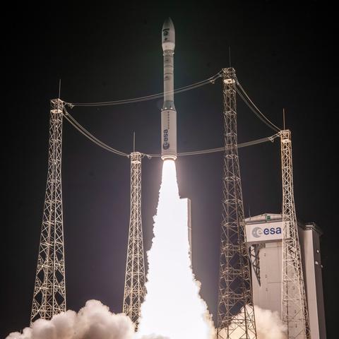The image shows a nighttime rocket launch with bright flames and smoke at the base as the rocket ascends. The white rocket features the ESA logo on its body, alongside smaller ones. The launchpad also features the same logo.