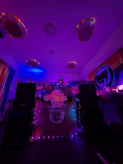 Me in a cat Onesy with a pride flag on the left under a lot of toiletry decoration in front. The room is covered in purple light