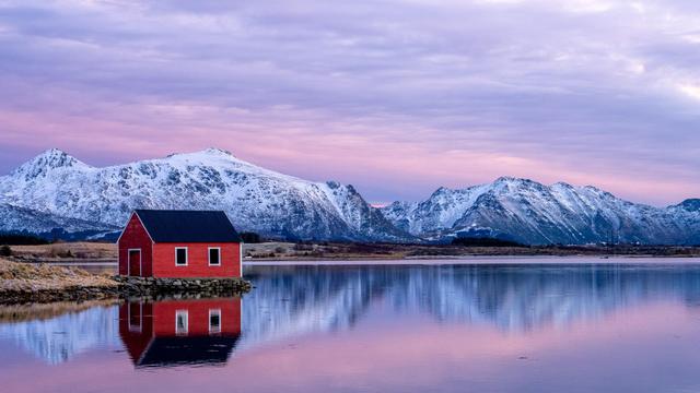 The image features a peaceful landscape with a red house situated by a calm lake. Behind the house are snow-covered mountains under a pink and purple sky, reflecting in the water. This creates a serene and picturesque setting.