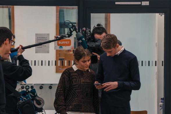 Teilnehmende der Jugendkonferenz prüfen auf einem Handy Aufnahmen, die gemacht wurden. Man sieht einen Kameramann, der ihnen dabei über die Schulter schaut und den Mikrofongalgen, den ein anderer Mann über ihre Köpfe hält.
