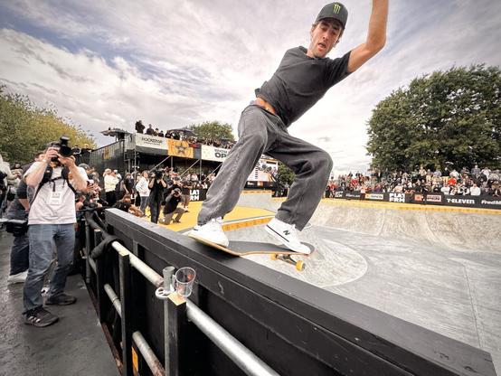 A male skateboarder performs a frontside tail stall on a bowl's extension on a mostly cloudy day.