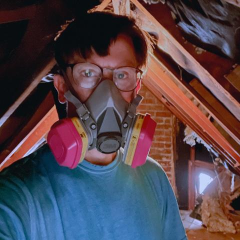 Me in a blue t-shirt with my face covered by a pink and gray, cartridge filtration mask while in an attic with flaking insulation.