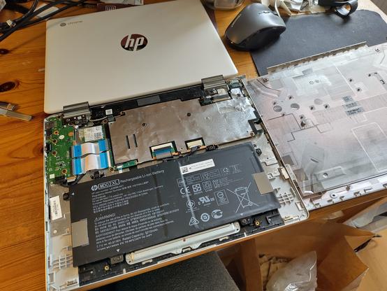 Geöffnete Unterseite eines HP-Chromebooks mit sichtbarer Hardware: der Akku, das Mainboard und andere Komponenten liegen frei. Der Laptop ruht auf einem Holztisch, daneben sind weitere Werkzeuge und Bauteile verstreut. Um den Schreibschutz der Firmware auszuschalten, muss das Mainboard vom Akku getrennt werden.