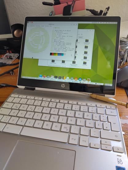 Ein auf einem Tisch liegendes silbernes HP Chromebook x360 Convertible, auf dem Ubuntu MATE ausgeführt wird. Auf dem Bildschirm ist ein Terminalfenster mit Systeminformationen zu sehen. Im Hintergrund sind verschiedene Kabel und Werkzeuge zu erkennen, auf der Tastatur liegt ein Stift.