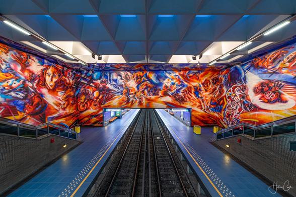 Wide angle symmetrical view from above on colourful fresco overhanging the platforms, at Hankar metro station in Brussels, at blue hour.  A dark blue sky is visible under a geometric glass structure in the ceiling, that reminds of the shapr of a snake face. Tiny black lamps hanging from the structure picture the snake's eyes. 
The wildly colorful “Notre Temps” fresco was painted  by Roger Somville, leader of the Belgian Expressionists Movement in 1976. The mural uses a lot of bright and strident colors in shades of orange and red to represent the struggles of the working class to attain social and economic  justice.