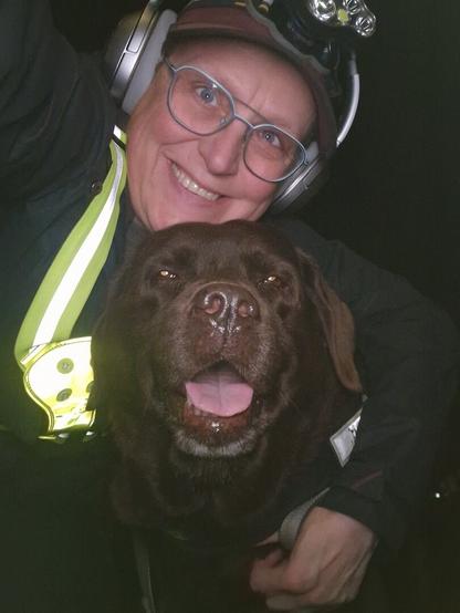 Night time selfie of a person and a chocolate brown Labrador with greying snout. The person is wearing a black jacket, yellow safety harness, red cap, silver headphones and blue-rimmed glasses. On their cap rests a headstrap torch in off position. Their free hand is around the dog's shoulders. Both are facing the camera. The dog's mouth is slightly open, showing off some blep. The person is smiling and enjoying the time with her furry love.