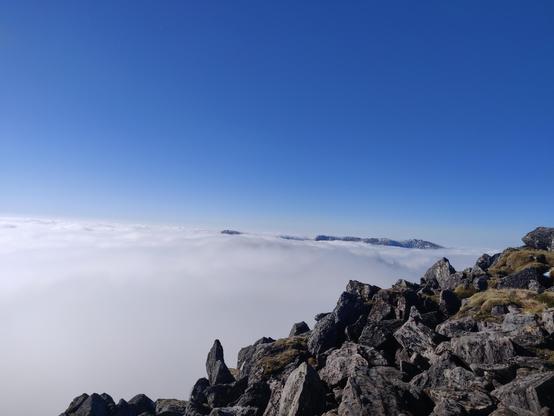 In the foreground, rocky terrain as you'd expect to find atop a mountain. Above, perfectly blue skies. Below, what looks like an undulating sea of cloud carrying on as far as the eye can see. In the distance, a range of peaks poke through the cloud looking like a distant island