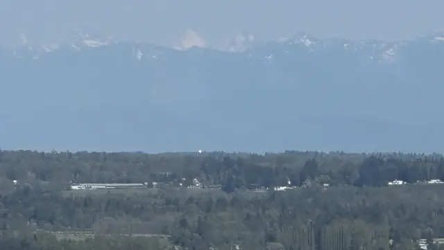 A zoomed in photo looking up a valley toward distant mountains. I white dot on a hill is seen poking above the trees, this is the weather radar.